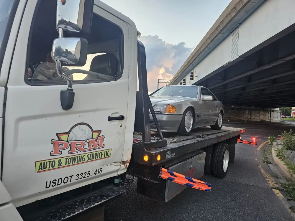 PRM Auto and Towing white flatbed tow truck carrying a silver convertible at dusk.