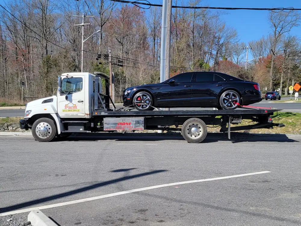 White PRM flatbed tow truck hauling a black Bentley sedan in side profile on a sunny day.