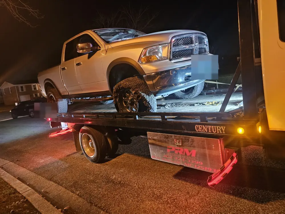 A PRM Auto and Towing flatbed carrying a lifted pickup truck at night, marker lights lit.